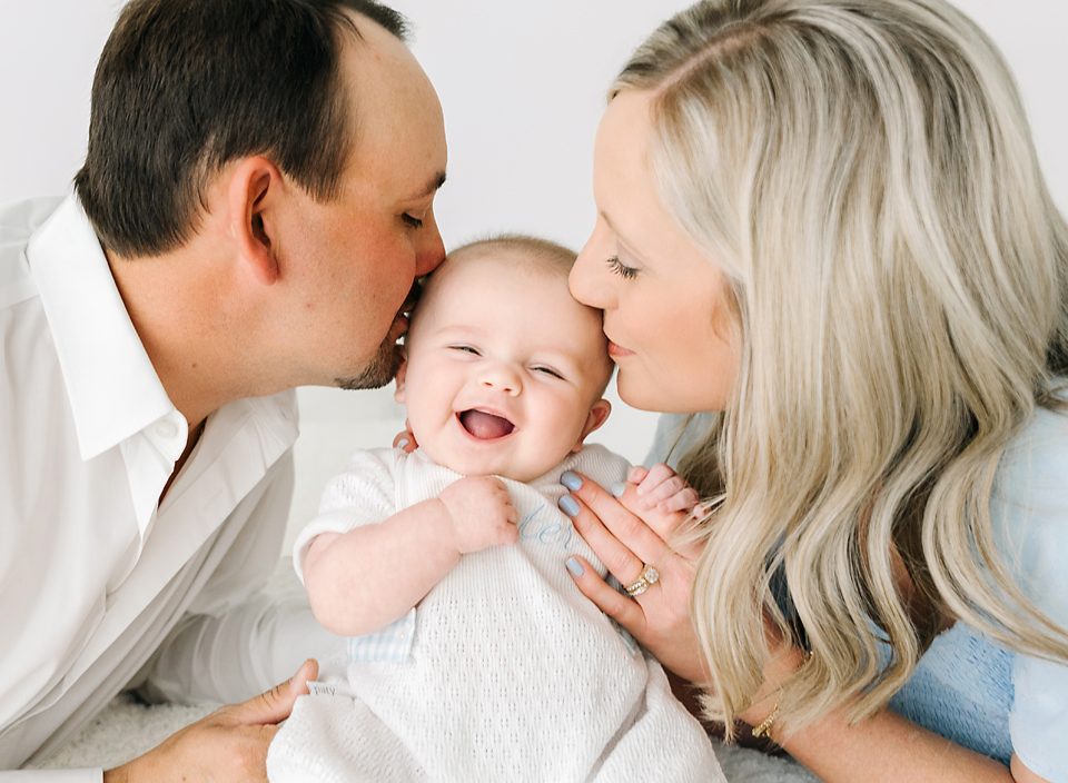 Mom and dad kissing 4 month old baby and baby is smiling big. Baby Studio Photography in Meridian, MS